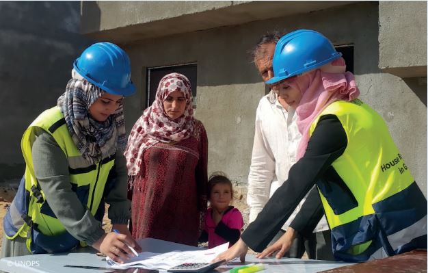 A male and female in a rural village are flanked by two woman in High visability vests and hard hats. They are  examining plans on an outdoor tableTwo wo