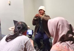 A person using sign language is being recorded by two other people. One person is filming with a smartphone on a tripod, and another person is watching while wearing a pink hijab.