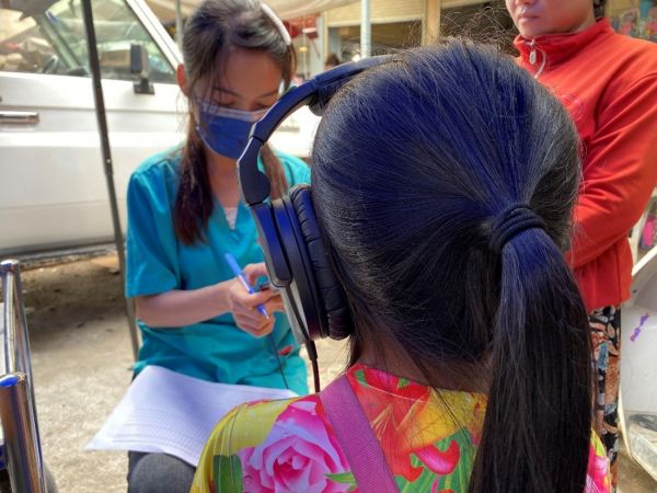 A woman in blue scrubs is sitting in front a young girl wihose dark hair is in a pony tail.  The young girl is wearing large headphones and has her back top the camera