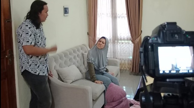 Three people are in a living room. The screen of  a video camera is in the corner of the image. A dark haired man leans against a pale wall he is talking to a woman sitting on a couch. A second woman sits on the titled floor. The women wear headscarfs