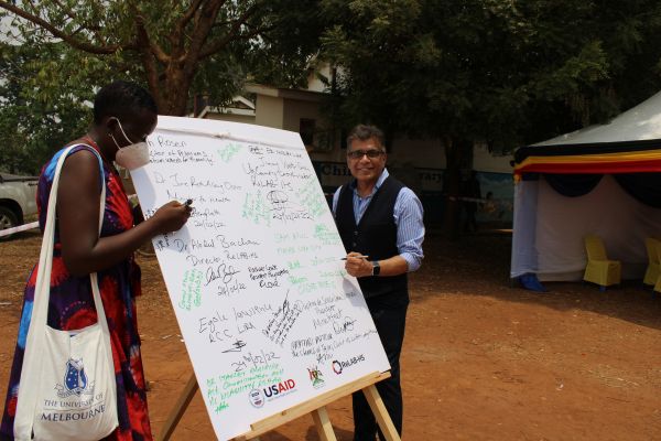 A Ugandan Woman about to sign a board with many signatures. A man a tsanding on the left about to sign