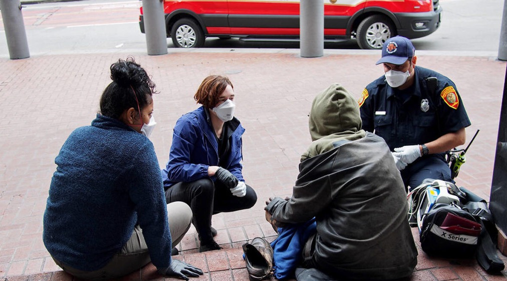 Two women and a Man in police uniform crouch respectfully in front of a homeless person.