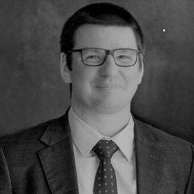A black and white head shot of  Mr John Demagistris.  He has short dark hair he is wearing glasses, and a suit and tie. 