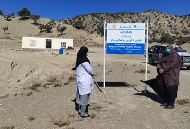 Two women are standing facing each other in front of a FHH sign in a remote area.  In the back ground is a portable building at the base of a sparsely vegetated hill..