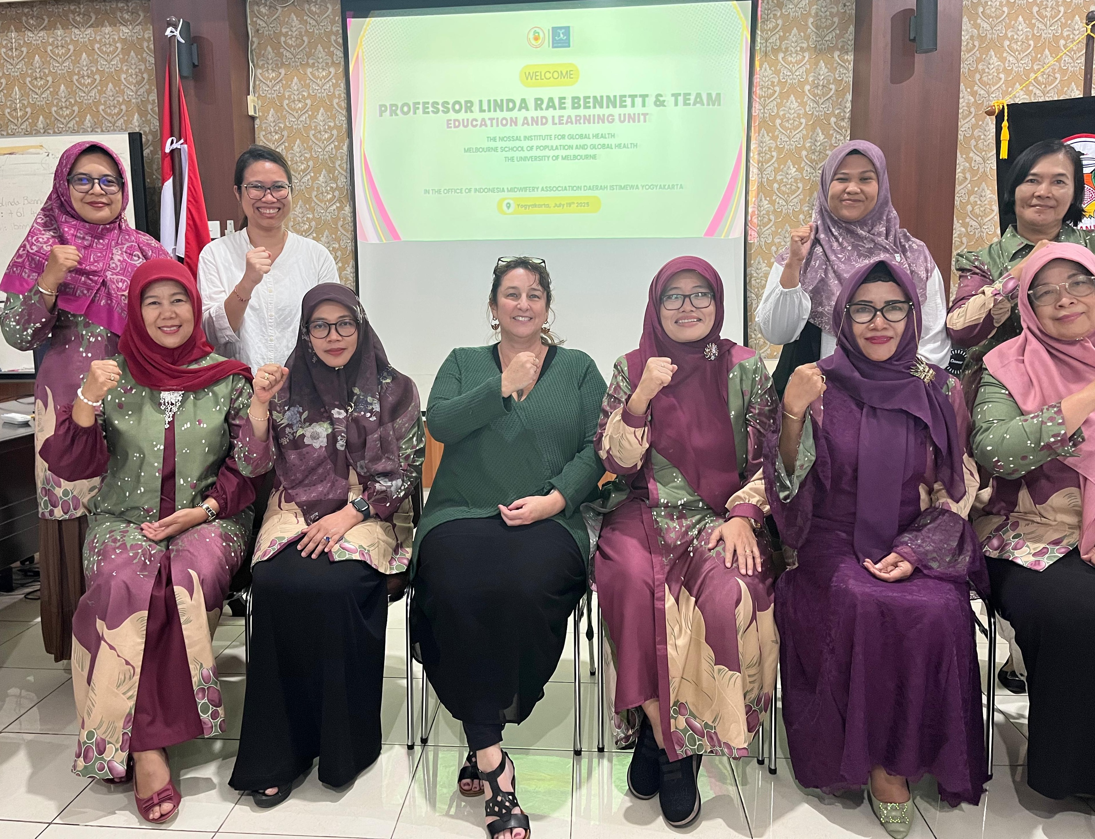 Professor Linda Rae Bennett, Mitha and UGM colleagues meeting with the Indonesian Midwives Association in Yogyakarta. 6 women are sitting 4 women stand behind. They are smiling but each also has a their right arm half raised with a fistsmiling for the c
