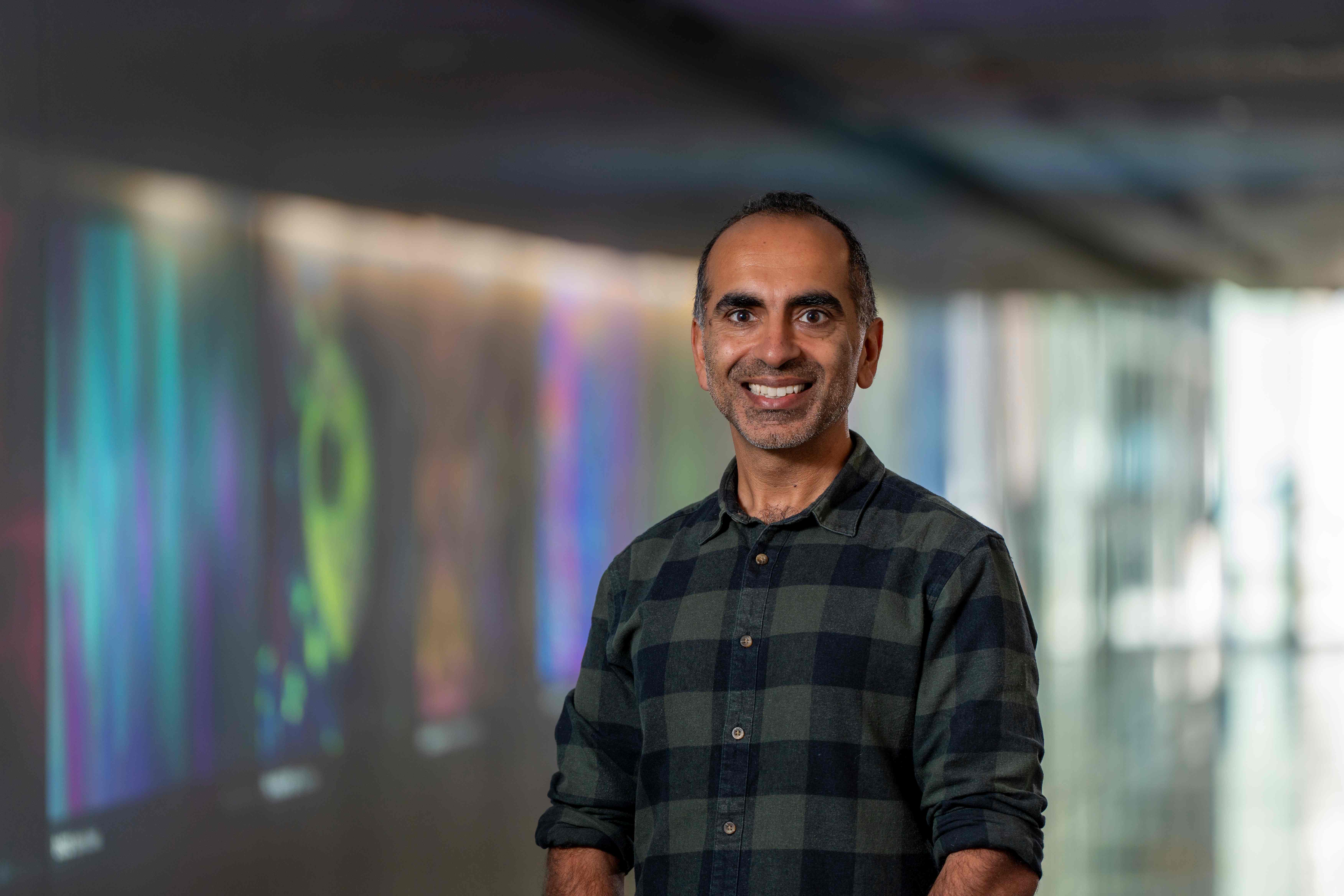 Professor Sant-Rayn Pasricha is smiling and standing in front of scientific artwork