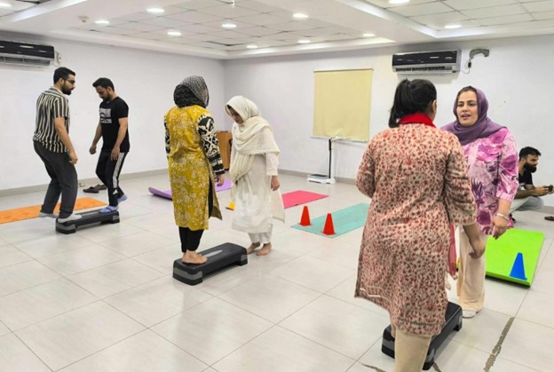 A room is set up with exercise stations. In the foreground are three set of two people.  Each pair is balancing at the ends of step ups. There are yoga mat is the background. The two pairs of woman are wearing shalwar kameez.