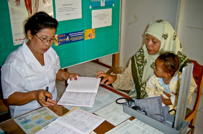 2 Indonesian women sit adjacent to each other at a wooden desk. One is a nurse the hiiab wearing patient is holding a baby. They a having a medical consultation
