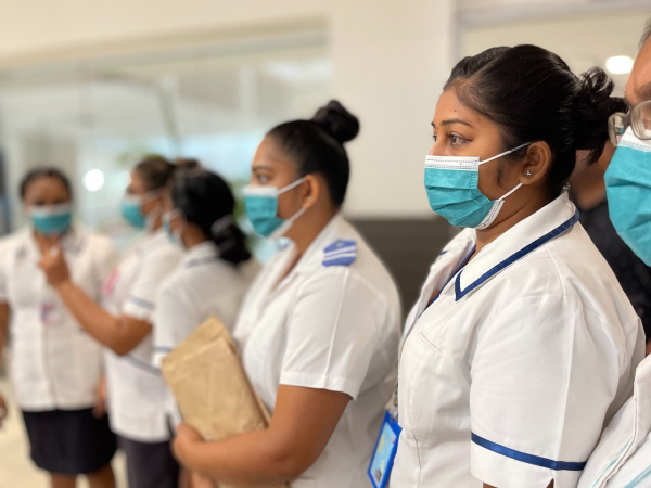 A group of Fijian female nurses are standinig paying atten to a speaker off camera. The vare in white uniformasd and wear blue surgical face masks. One womans is in focus the other are progresivley blurred. Image Curtsey of WHO Western Region