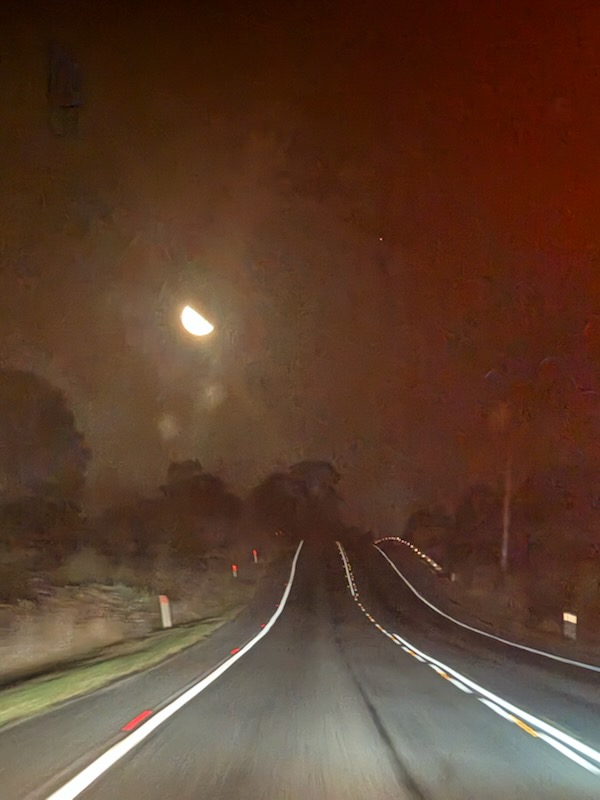 Photograph of the view down a highway at night, slightly blurred. The straight road disappears over a small hill, and the moon is visible overhead. Trees and grass line the road.  