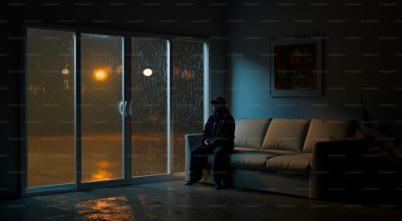Image of someone sitting alone in a dark room, looking out through the window. They are isolated. 