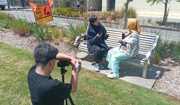Two people are sitting on a parke bench they are having a conversation using sign language. They are being filmed by someone using a small camera on a tripod.